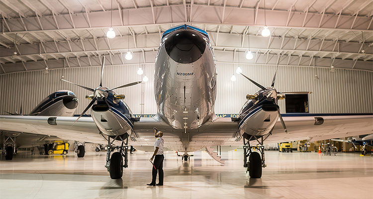 A vintage aircraft is parked in a spacious hangar, with a person standing in front of it