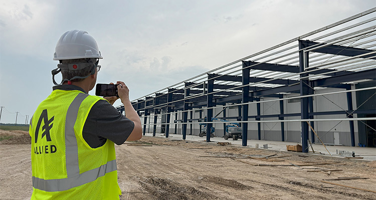 Allied Steel Buildings leads as the partner of choice for the world’s toughest structural steel projects a construction worker, wearing a hard hat and high-visibility vest with "ALLIED" written on it, taking a photo or video of a building under construction