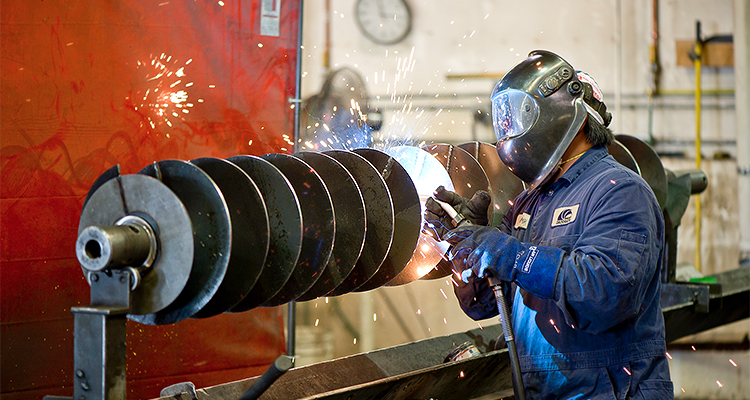 a worker performing industrial welding on a conveyor screw component.