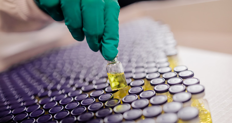 a person wearing a green protective glove holding a small glass autosampler vial filled with a yellowish liquid