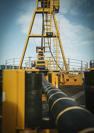 A long, thick black pipe or riser extends toward a large yellow mechanical tower.