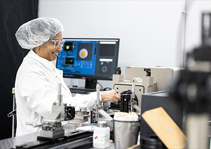 a female researcher wearing a white lab coat, a hair net, and glasses, smiling while working in a laboratory
