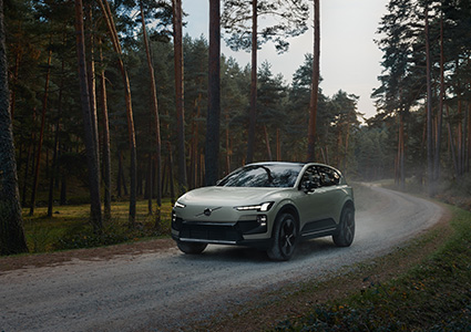 a light-colored, all-electric Volvo EX60 mid-size SUV driving on a dirt road through a dense forest