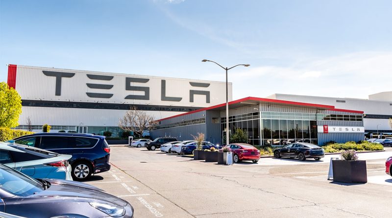 Tesla manufacturing facility exterior with large TESLA signage, parked cars in the foreground, and a modern glass-fronted entrance under a clear blue sky.
