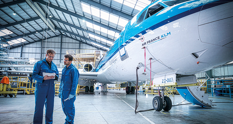 two aircraft maintenance engineers in blue jumpsuits conversing in a large, well-lit hangar