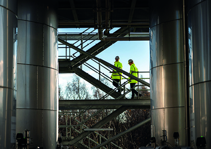 featuring workers on a metal staircase between large stainless steel storage tanks