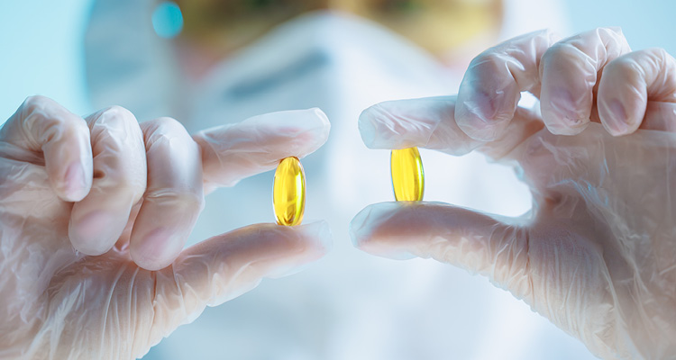 a person wearing a white lab coat, a face mask, safety goggles, and translucent disposable gloves holding two translucent, yellow oval softgel capsules