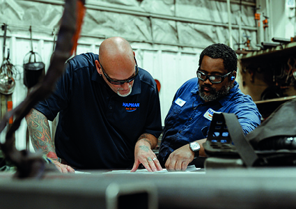 two people wearing safety glasses and work shirts while examining blueprints or technical documents in an industrial or workshop environment