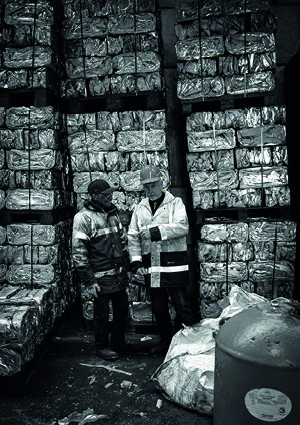 two workers standing in what appears to be a recycling or industrial facility, surrounded by large, rectangular bales of compressed materials