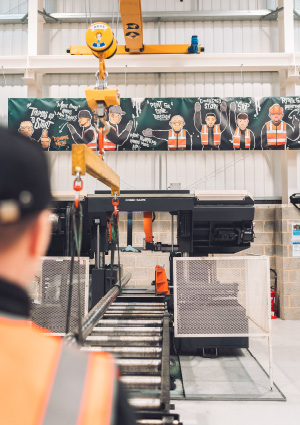 an over-the-shoulder view of a worker monitoring a large horizontal band saw and an overhead hoist system inside an industrial workshop