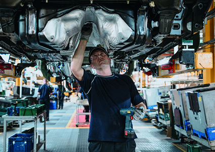 an assembly line worker performing manual installation tasks underneath a vehicle chassis on an automotive production floor