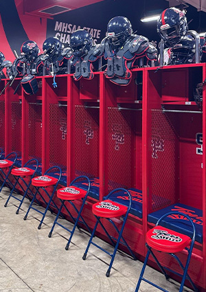 a row of red football lockers outfitted with matching shoulder pads, helmets on top, and blue-and-red folding stools lined up in front
