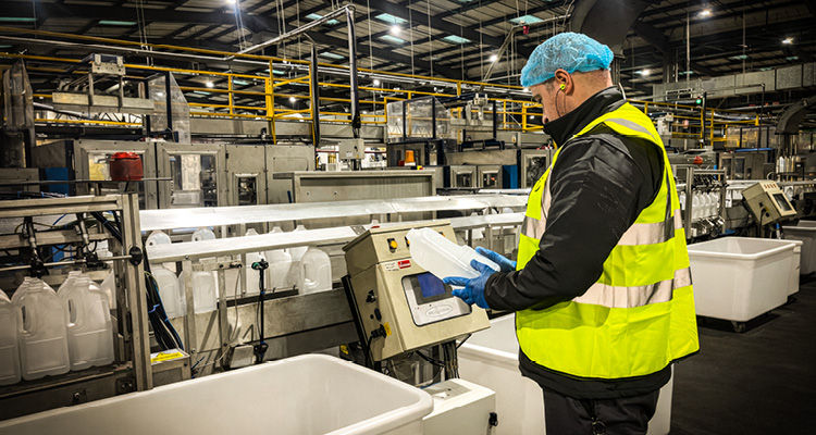 a worker in a high-visibility vest and hairnet inspecting a plastic container on a bottling or packaging assembly line
