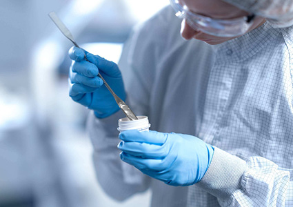 a laboratory worker wearing blue protective gloves and safety gear while opening or sampling from a small white container with a spatula