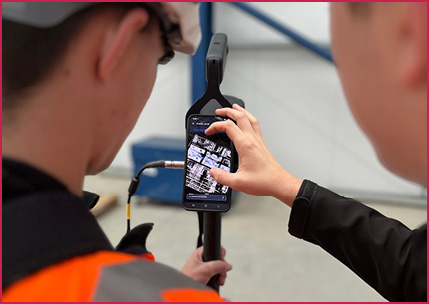 two individuals in an industrial or construction setting interacting with a smartphone mounted on a pole-like device