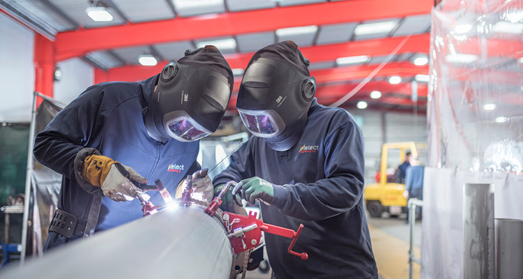two industrial workers wearing protective welding helmets while fabricating large metal pipework in a workshop