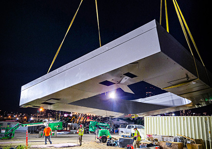 a large, rectangular precast concrete or steel structural module being hoisted into the air by heavy-duty cranes