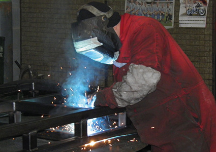 a person wearing a protective welding helmet and a red jumpsuit while arc welding a metal frame