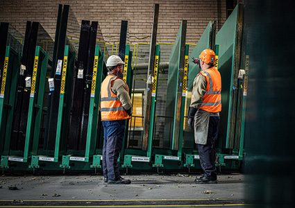 two workers in a manufacturing warehouse inspecting large panes of architectural glass stored on green stillage racks