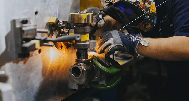 a worker wearing a protective welding helmet and gloves while precision welding metal pipe components